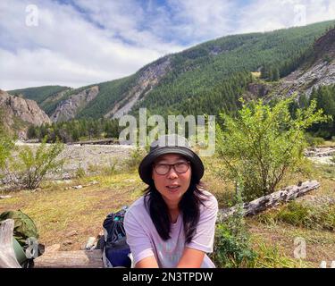 Ein asiatisches Mädchen mit Brille und Hut macht ein Selfie vor dem Hintergrund des Himmels und der Berge in Altai. Stockfoto