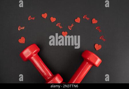 Gym-Kurzhanteln und rote Dekorationen in Herzform. Fitness-Liebesgeschenk zum Valentinstag, Heiratsantrag, Verlobung, Jahrestag, Hochzeit. Stockfoto