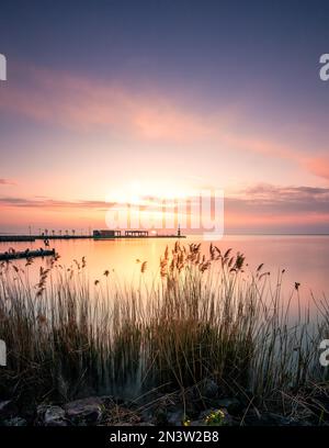 Ufer an einem See, mit Schilf. Im Hintergrund befindet sich eine Bootsanlegestelle und die Sonne geht auf, tihany, Balaton, Plattensee, Ungarn Stockfoto