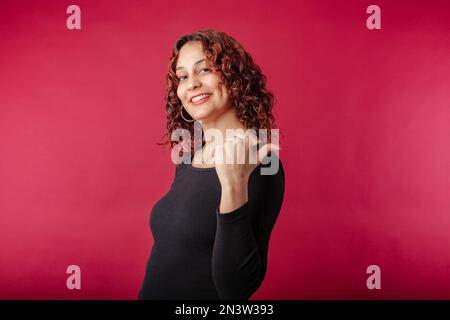 Junge Rothaarige in schwarzem, geripptem Kleid, isoliert auf rotem Hintergrund und zeigt mit dem Daumen nach hinten auf den Kopierbereich des Arbeitsbereichs. Ich schaue mich um Stockfoto