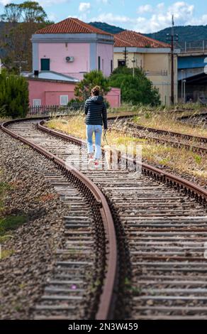 Junge Frau mit Hund, die auf dem stillgelegten Bahnbett in der Stadt Figari, Golfo Aranci, Sardinien, Italien spaziert Stockfoto