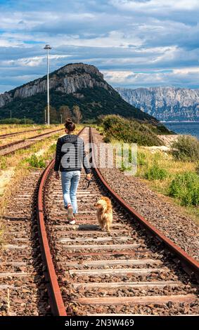 Junge Frau mit Hund, die auf dem stillgelegten Bahnbett in der Stadt Figari, Golfo Aranci, Sardinien, Italien spaziert Stockfoto