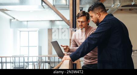 Geschäftsmann, der ein Technologieprojekt mit seinem Kollegen in einem Büro bespricht. Zwei Geschäftsleute, die einen Laptop auf einem Balkon benutzen. Männliche Profis Stockfoto