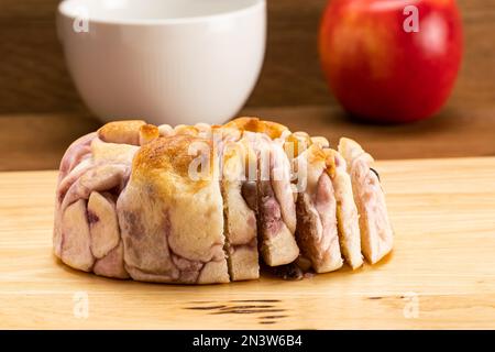 Seitlicher Blick ein Laib aus lila Süßkartoffelbrot mit gemischten Bohnen auf Holzbrett, einer weißen Keramiktasse Kaffee und einem reifen roten Apfel Stockfoto