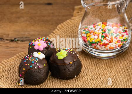 Schokoladenkugeln mit bunten Regenbogenstreuseln und Zuckerblume auf Sacktuch und bunten Regenbogenstreuseln in durchsichtigem Glaskrug Stockfoto