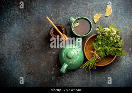 Kräutertee mit Kamille, frische medizinische Kräuter und Blumen, Teetasse, Teekanne, Glas Honig, Zitrone Draufsicht auf Stein Hintergrund, Kräuteralternative Stockfoto