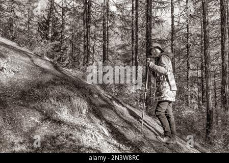 Ein Schwarz-Weiß-Foto einer asiatischen Travelerin mit Brille und Hut steht vor einem steilen Aufstieg auf einem Berg mit Wanderstöcken. Stockfoto