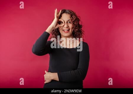 Glückliche Frau in schwarzem Kleid, isoliert auf rotem Hintergrund, mit Blick durch das Okey-Schild. Glücklich, selbstbewusst und alles ist in Ordnung. Gesichtsausdruck Stockfoto