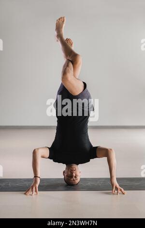 Ein Mann führt im Fitnessstudio eine Yoga-Pose mit Unterstützung auf dem Kopf durch. Yogi im Kopfständer. Stockfoto