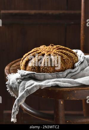 Stillleben mit frisch gebackenem, leckerem runden Brot aus Gerste und Maismehl mit einer Tischdecke aus Naturleinen auf einem alten Holzstuhl. Dunkler rustikaler Stil. Stockfoto