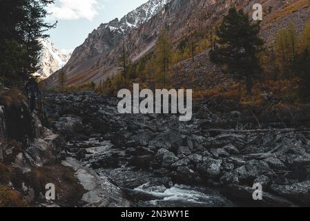 Ein Mädchen mit Wanderstöcken klettert im Schatten auf einem steilen Pfad vor dem Hintergrund eines Flusses und Berge mit Gletschern und Sno Stockfoto