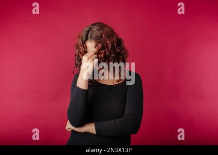 Porträt einer jungen rothaarigen Frau, die ein isoliertes Kleid auf rotem Hintergrund trägt, müde, reibende Nase und Augen, Müdigkeit und Kopfschmerzen. Stress und Frustr Stockfoto