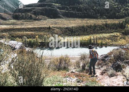 Ein Mädchenreisender erklingt einen steilen Pfad vor der Kulisse eines Flusses und der Berge in Altai. Stockfoto