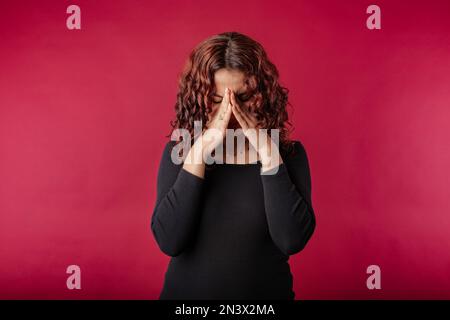 Rothaarige Frau, die isoliert auf rotem Hintergrund steht, so müde vom Leben. Schmerzen in den Nebenhöhlen. Schmerz mit Kopfschmerzen, Augenbelastung emotionaler Stress Stockfoto