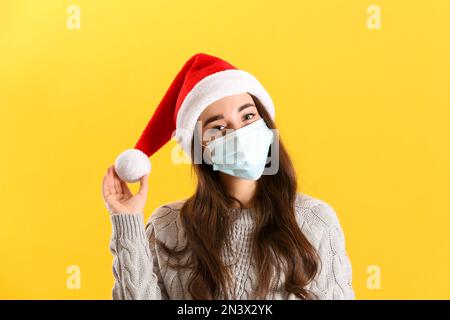 Wunderschöne Frau trägt Weihnachtsmann Hut und medizinische Maske auf gelbem Hintergrund Stockfoto