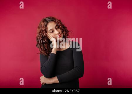 Eine junge weiße Frau, die ein Kleid trägt, das isoliert auf rotem Hintergrund steht, neigt sich den Kopf auf die Handfläche und sieht pessimistisch auf den leeren Kopierraum. Nicht ha Stockfoto