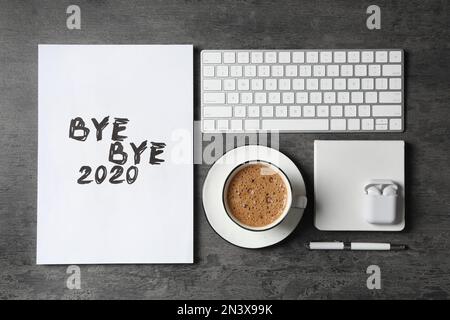 Weißes Blatt Papier mit der Phrase „Bye Bye 2020“ neben Kaffee, Tastatur und kabellosen Ohrhörern auf grauem Tisch, flach liegend Stockfoto