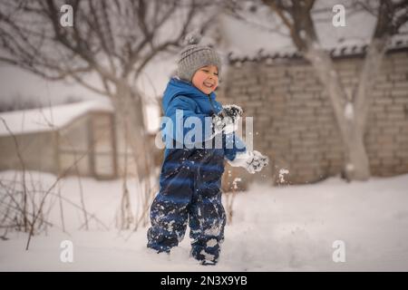 Lustiger kleiner Junge in blauer Winterkleidung läuft während eines Schneefalls. Winteraktivitäten im Freien für Kinder. Süßes Kind mit warmem Hut Stockfoto