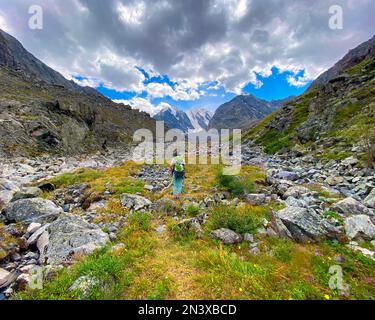 Ein Touristenmädchen mit Rucksack geht auf einem steinernen Pfad im Altai-Gebirge vor dem Hintergrund von Gletschern und Schnee im Sommer spazieren. Stockfoto