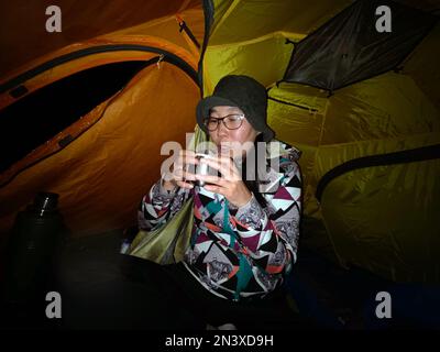 Eine asiatische Reisende sitzt nachts in einem Zelt und trinkt Tee. Stockfoto