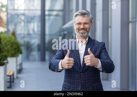 Porträt eines erwachsenen Investors außerhalb des Bürogebäudes, ein leitender Mann lächelt und schaut in die Kamera und zeigt Daumen nach oben. Stockfoto