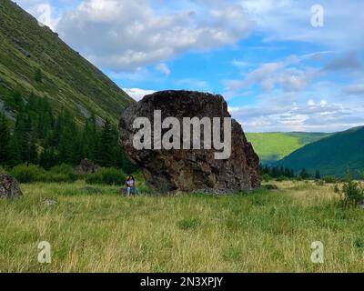 Ein Touristenmädchen mit einem Rucksack mit Brille und einem Hut steht auf Trekkingstöcken vor dem Hintergrund eines großen Steins im Altai-Gebirge. Stockfoto