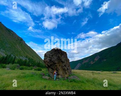 Eine Reisende mit einem Rucksack in Brille und Hut steht auf Trekkingstöcken vor dem Hintergrund eines großen Steins im Altai-Gebirge Stockfoto