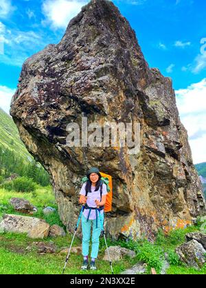 Eine Reisende mit einem Rucksack in Brille und Hut steht auf Trekkingstöcken vor dem Hintergrund eines großen Steins im Altai-Gebirge. V Stockfoto