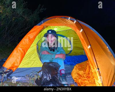 Eine asiatische Reisende mit Laterne sitzt nachts in einem Zelt und lächelt. Stockfoto