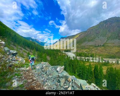 Weibliche Reisende mit Rucksack und Wanderstöcken erklimmen im Sommer einen steilen Steinpfad in den Bergen in der Nähe des Flusses in Altai. Stockfoto