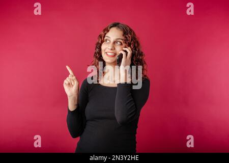 Glückliche Frau, die isoliert auf rotem Hintergrund steht und am Telefon spricht, sich den Kopierbereich ansieht und mit dem Zeigefinger zeigt. Anzeige einer Anzeige Stockfoto