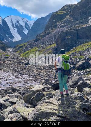 Ein Mädchen mit Rucksack wandert auf einem steinernen Pfad im Altai-Gebirge vor dem Hintergrund von Gletschern und Schnee im Sommer. Ver Stockfoto