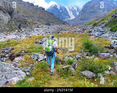 Ein Mädchen mit Rucksack wandert auf einem Bergweg durch die Berge und Felsen von Altai vor dem Hintergrund von Gletschern und Schnee im Sommer. Stockfoto