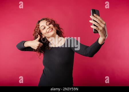 Süße weiße Frau, die ein schwarzes geripptes Kleid trägt, isoliert auf rotem Hintergrund, Selfie macht und Gesten wie Daumen hoch zum Smartphone macht, kommuniziert Stockfoto