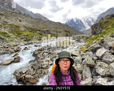 Ein lächelndes asiatisches Mädchen mit Brille und Hut macht am Telefon ein Selfie vor dem Hintergrund von Gletschern und schneebedeckten Bergen in der Nähe eines Bachs in Al Stockfoto