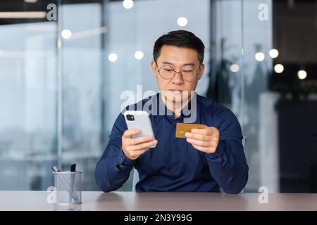 Ein seriöser und denkender asiatischer Mann im Büro, der die App auf dem Telefon und der Bankkreditkarte für den Kauf und die Buchung von Online-Shops verwendet, konzentrierte sich auf die Eingabe von Passwörtern am Arbeitsplatz. Stockfoto