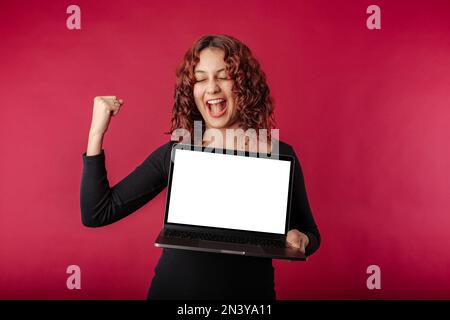 Die süße Millennials in einem schwarzen Rippenkleid, die isoliert auf rotem Hintergrund mit geschlossenen Augen steht, hält ein Notebook in der Hand und ist unglaublich Stockfoto