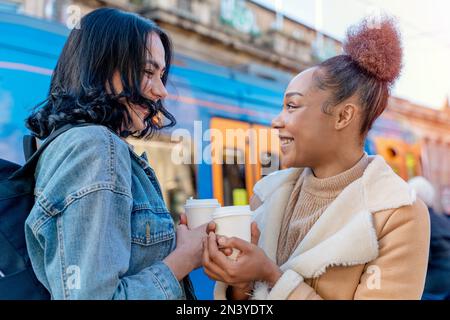 Zwei Frauen in einer Jeansjacke reden miteinander, trinken Kaffee und warten auf eine Straßenbahn an der Haltestelle Lifestyle Foto Stockfoto