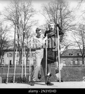 1950er Paar. Die Schauspielerin Annalisa Ericson trainiert, um zu lernen, wie man auf Stelzen geht und das Gleichgewicht hält. Sie bekommt Hilfe von einem Mann, der sie im Gleichgewicht hält, damit sie nicht fällt. Obwohl sie so hilfreich war, zeigt ihr Gesichtsausdruck, dass sie nicht wirklich gelernt hat, wie. Schweden 1956 Kristoffersson Ref BX64-3 Stockfoto