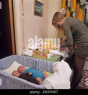 1970er Mutter. Eine Frau putzt und wechselt Windeln auf ihren drei Babys, die auf einem Wickeltisch liegen. Sie hat Drillinge und zwei sind im Bett. Schweden 1972 Ref. BV105-3 Stockfoto