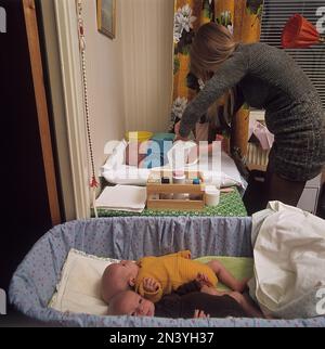 1970er Mutter. Eine Frau putzt und wechselt Windeln auf ihren drei Babys, die auf einem Wickeltisch liegen. Sie hat Drillinge und zwei sind im Bett. Schweden 1972 Ref. BV105-4 Stockfoto