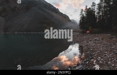 Ein weiblicher Fischer steht mit einer Angelrute in der Nähe eines hohen türkisfarbenen Sees mit einem Spiegelbild von Shavlinskoye neben einem Zelt im Wald und smok Stockfoto