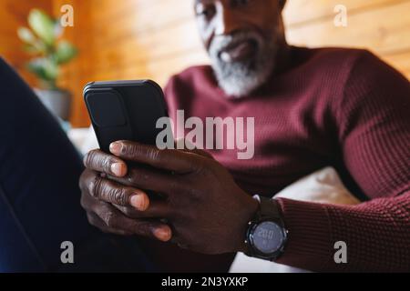 Nahaufnahme eines afroamerikanischen Seniorenmanns, der sein Smartphone benutzte, während er auf der Couch in der Blockhütte saß Stockfoto