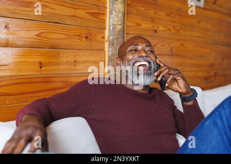 Glatzköpfiger, fröhlicher afroamerikanischer Senior-Mann, der sich über Handy unterhält, während er auf der Couch in der Blockhütte sitzt Stockfoto