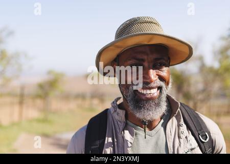 Porträt eines lächelnden, bärtigen afroamerikanischen Seniorenmanns mit Hut, der im Wald unter klarem Himmel wandert Stockfoto