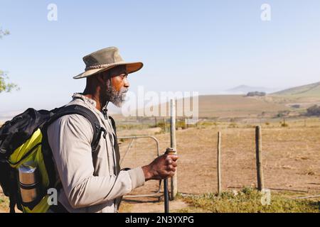 afroamerikanischer Seniorist mit Rucksack und Wanderstöcken, der an Land vor klarem Himmel spaziert Stockfoto