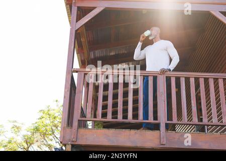 Blick aus dem niedrigen Winkel auf einen afroamerikanischen Senioren, der Kaffee trinkt, während er auf dem Balkon am Geländer steht Stockfoto