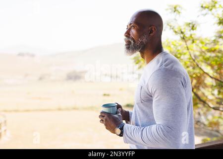 Kahlköpfiger afroamerikanischer Seniorenmann mit Tasse, der sich Gedanken macht und auf dem Balkon vor klarem Himmel steht Stockfoto