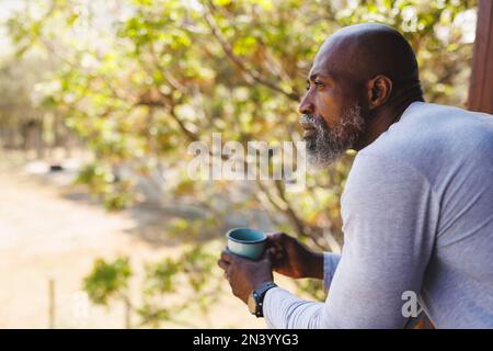 Seitenansicht eines aufmerksamen, kahlköpfigen, afroamerikanischen Seniorenmanns mit Kaffeetasse auf dem Balkon Stockfoto