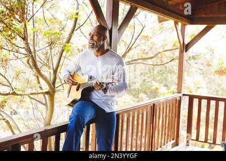 Kahlköpfiger afroamerikanischer Senior-Mann, der Gitarre spielt und singt, während er auf dem Geländer auf dem Balkon sitzt Stockfoto